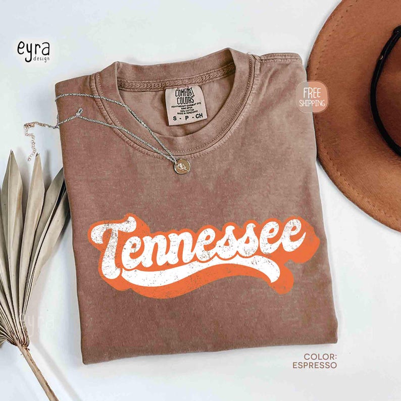 May include: Espresso-coloured t-shirt featuring the word "Tennessee" in white with an orange outline. The shirt has a crew neck and short sleeves. A silver necklace with a small pendant rests on the shirt. The shirt is labelled "Comfort Colors" and is size S-P-CH.