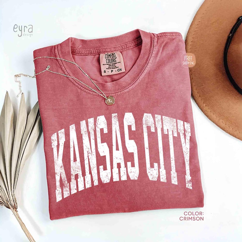 May include: Crimson-colored t-shirt with the text "KANSAS CITY" in distressed white lettering. A gold necklace with a pendant rests on the shirt. The shirt is a Comfort Colors brand, size S-P-CH.