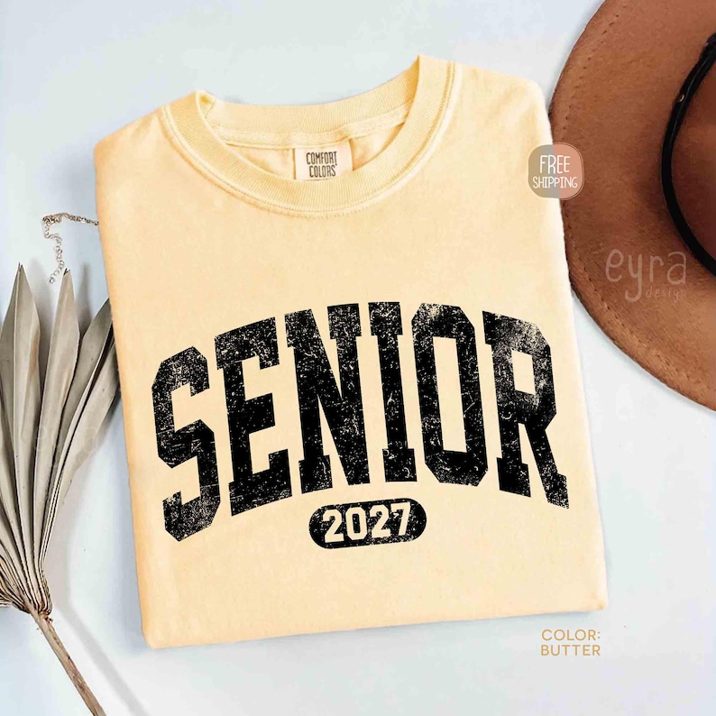 May include: A light yellow t-shirt with the word "SENIOR" in distressed black lettering arched across the front, with "2027" below. The shirt is a Comfort Colors brand.