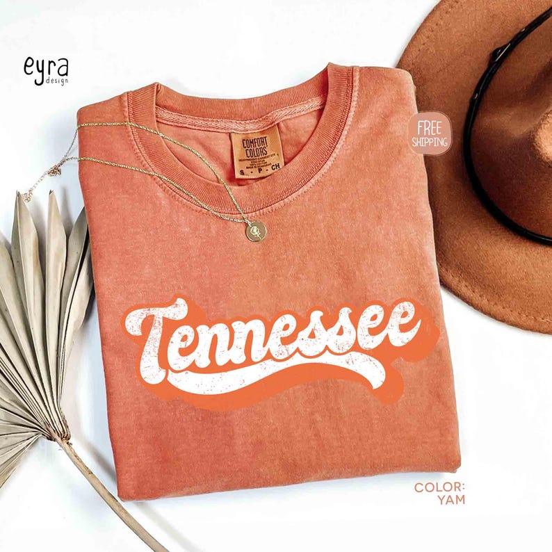 May include: A yam-coloured t-shirt with the word "Tennessee" in white, retro-style lettering. The shirt has a crew neck and a small tag. A gold necklace and a brown hat are also visible.
