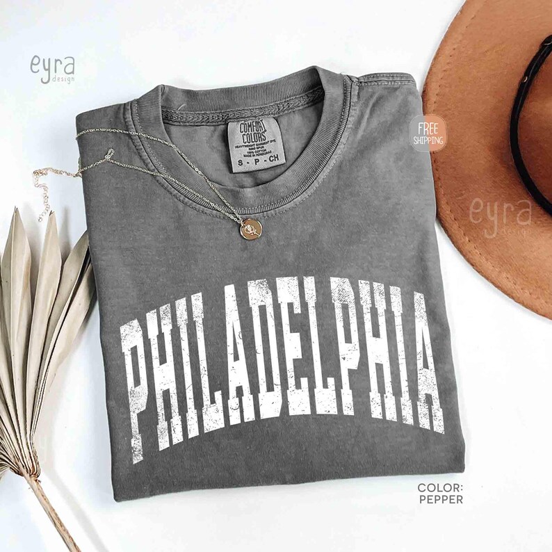 May include: A charcoal grey short-sleeve t-shirt with a crew neckline featuring the word PHILADELPHIA printed in a large, distressed white arched font across the chest. The shirt includes a Comfort Colors brand label at the inner neck.