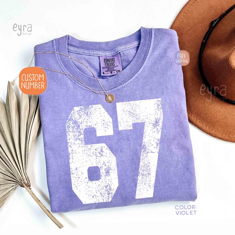 K&ouml;nnte beinhalten: Ein violettes Comfort Colors T-Shirt mit der Nummer 67 in wei&szlig;er, verblichener Schrift. Eine goldene Halskette und ein brauner Hut liegen neben dem Shirt. Das Shirt hat ein Etikett mit der Aufschrift "Comfort Colors". Die Farbe des Shirts ist "Violett".