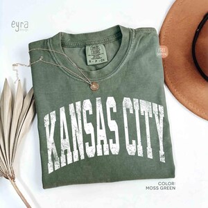 May include: Moss green t-shirt with the text "KANSAS CITY" in white, distressed lettering. A gold necklace with a pendant rests on the shirt. A brown hat is visible in the background.