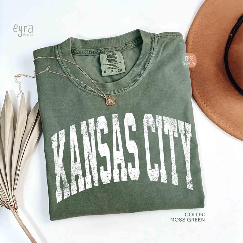 May include: Moss green t-shirt with the text "KANSAS CITY" in white, distressed lettering. A gold necklace with a pendant rests on the shirt. A brown hat is visible in the background.