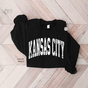 May include: Black sweatshirt featuring the text "KANSAS CITY" in white, arched lettering. The long-sleeved top has a crew neck and is made of a soft material. The color of the sweatshirt is black.