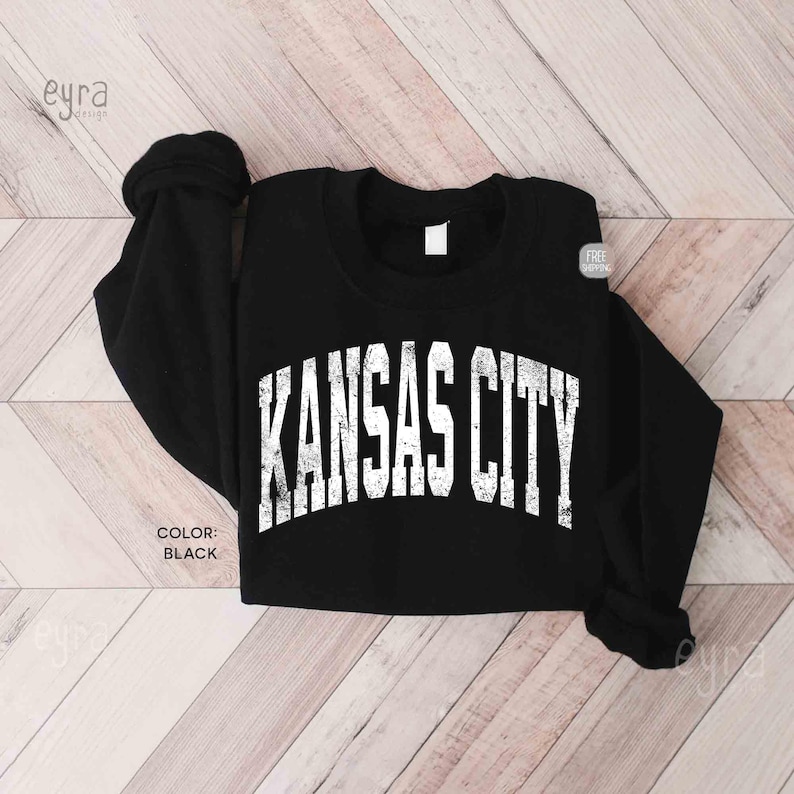May include: Black sweatshirt featuring the text "KANSAS CITY" in white, arched lettering. The long-sleeved top has a crew neck and is made of a soft material. The color of the sweatshirt is black.
