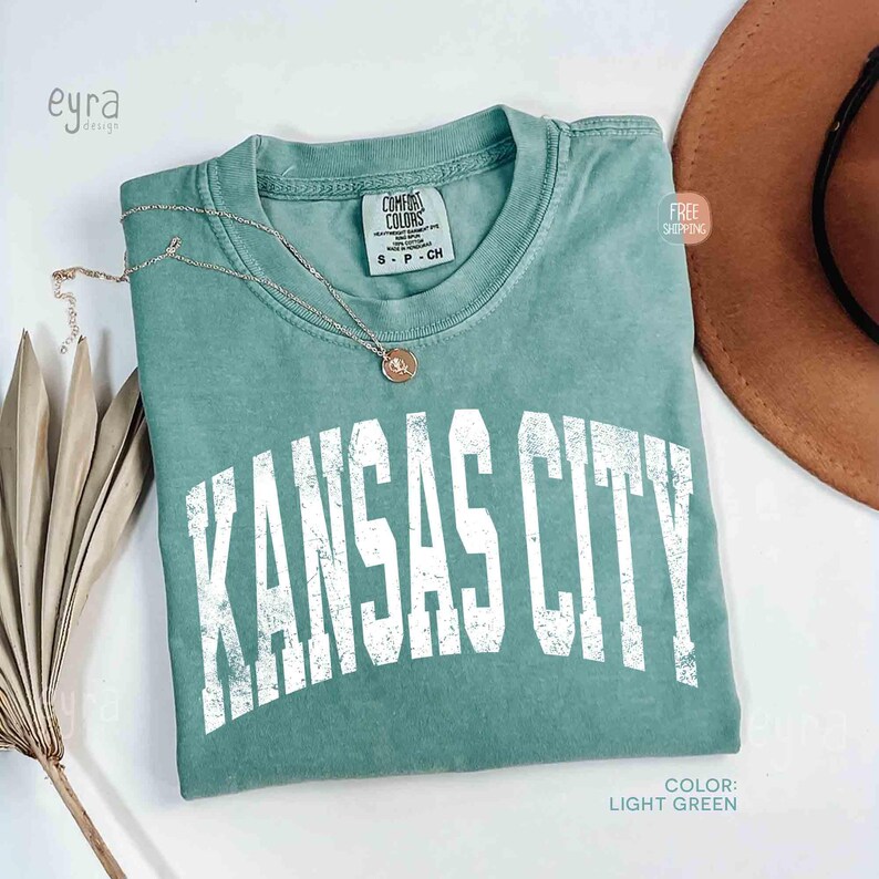 May include: Light green t-shirt with the text "KANSAS CITY" in white, distressed lettering. A gold necklace with a pendant rests on the shirt. The shirt is labeled "Comfort Colors" and is size S-P-CH.