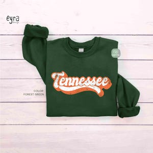K&ouml;nnte beinhalten: Ein lang&auml;rmeliges Sweatshirt in Waldgr&uuml;n mit dem Wort Tennessee, das in einer wei&szlig;en, abgenutzten Schreibschrift mit orangefarbenem Umriss mittig auf der Brust aufgedruckt ist. Das Kleidungsst&uuml;ck hat einen Rundhalsausschnitt und gerippte B&uuml;ndchen.