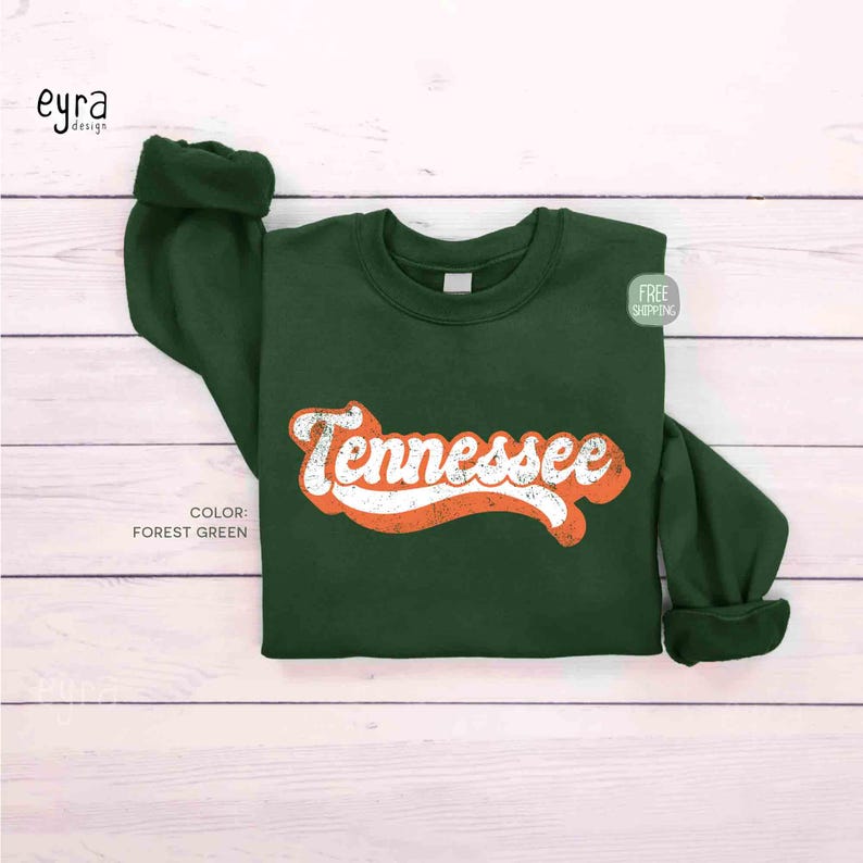 K&ouml;nnte beinhalten: Ein lang&auml;rmeliges Sweatshirt in Waldgr&uuml;n mit dem Wort Tennessee, das in einer wei&szlig;en, abgenutzten Schreibschrift mit orangefarbenem Umriss mittig auf der Brust aufgedruckt ist. Das Kleidungsst&uuml;ck hat einen Rundhalsausschnitt und gerippte B&uuml;ndchen.