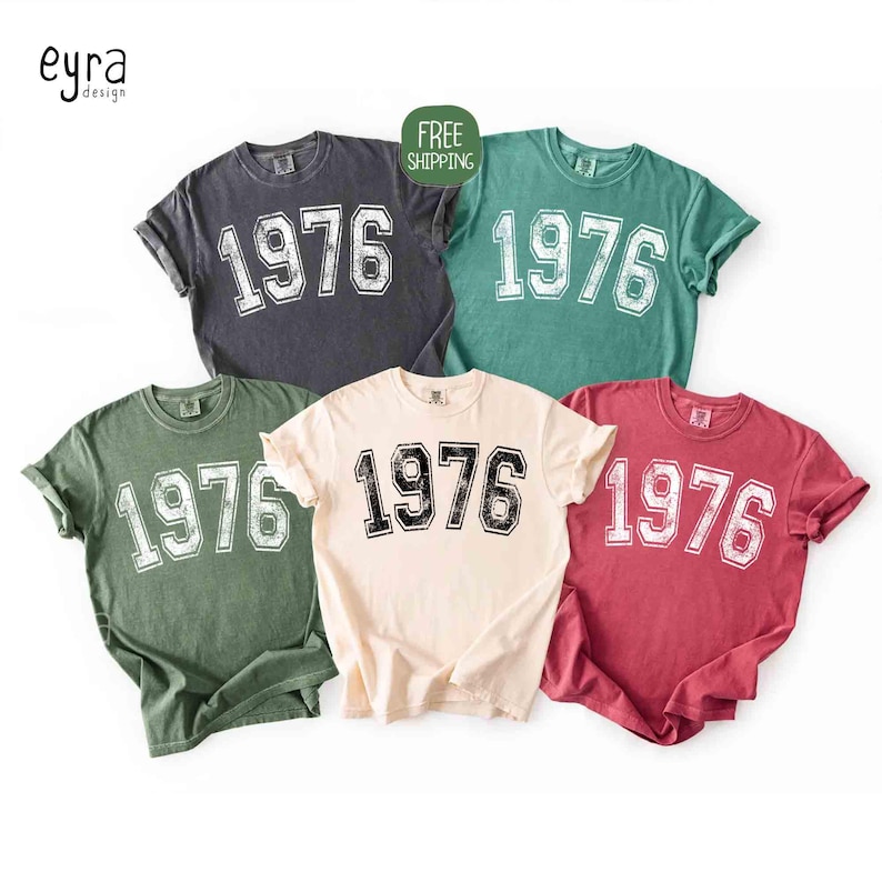 May include: Five short-sleeved t-shirts in various colors: charcoal, teal, olive green, cream, and maroon. Each shirt features the number "1976" in a distressed, collegiate-style font. The shirts are from "eyra design".