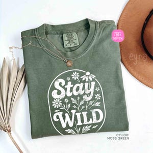 May include: A moss green t-shirt with the words "Stay Wild" in white script and floral design. A gold necklace with a pendant rests on the shirt. The shirt is folded on a white surface.