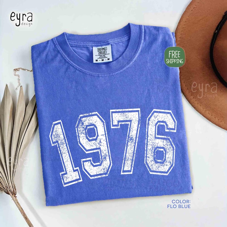 May include: A blue t-shirt with the number "1976" printed in white, distressed lettering. The shirt is folded and has a crew neck. A brown hat is partially visible in the background. The shirt is labeled "Flo Blue".