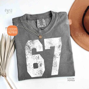 K&ouml;nnte beinhalten: Ein graues Comfort Colors T-Shirt mit der Nummer 67 in Wei&szlig;. Das Shirt hat einen Rundhalsausschnitt und kurze &Auml;rmel. Eine goldene Halskette mit Anh&auml;nger liegt auf dem Shirt. Die Farbe ist Pfeffer.