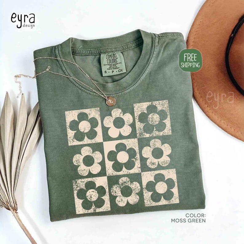 May include: Moss green t-shirt with a retro floral design. The shirt features a grid of nine flowers in a cream color. A silver necklace with a pendant is on the shirt. The shirt is labeled "Comfort Colors".