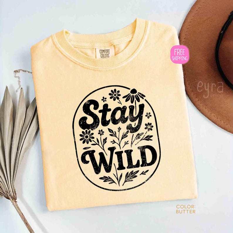 May include: A light yellow t-shirt with a crew neck. The shirt features a black oval design with the words "Stay Wild" and floral accents. The shirt is labeled "Comfort Colors" and the color is "Butter".