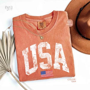 Puede incluir: Una camiseta de color coral con la palabra "USA" en letras blancas grandes y desgastadas y una peque&ntilde;a bandera americana. La camiseta tiene cuello redondo y una etiqueta que dice "Comfort Colors". Tambi&eacute;n se ven un collar dorado y un sombrero marr&oacute;n.