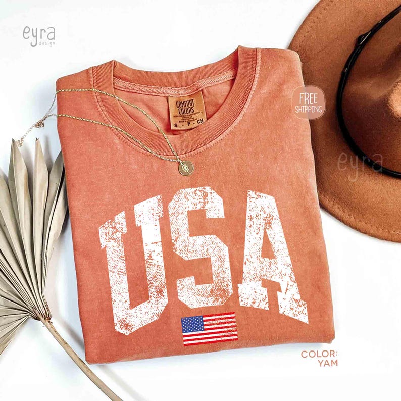 Puede incluir: Una camiseta de color coral con la palabra "USA" en letras blancas grandes y desgastadas y una peque&ntilde;a bandera americana. La camiseta tiene cuello redondo y una etiqueta que dice "Comfort Colors". Tambi&eacute;n se ven un collar dorado y un sombrero marr&oacute;n.