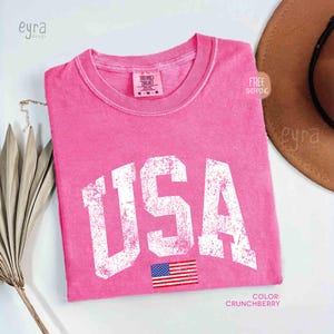 Puede incluir: Una camiseta rosa con la palabra "USA" en letras blancas desgastadas y una peque&ntilde;a bandera estadounidense. La camiseta tiene cuello redondo y mangas cortas. El color es "Crunchberry".