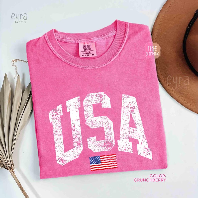 Puede incluir: Una camiseta rosa con la palabra "USA" en letras blancas desgastadas y una peque&ntilde;a bandera estadounidense. La camiseta tiene cuello redondo y mangas cortas. El color es "Crunchberry".