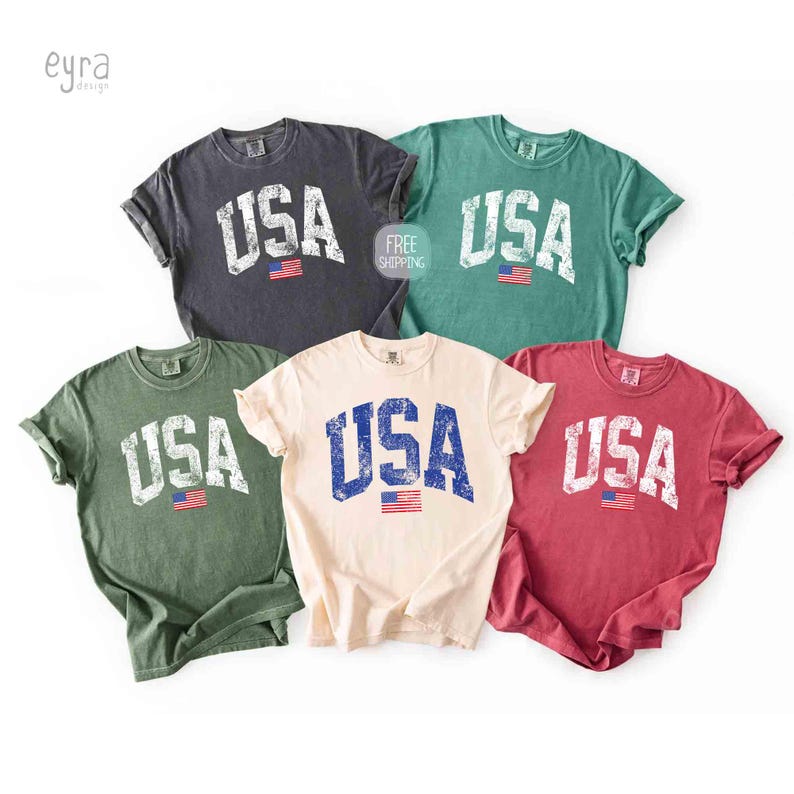 Puede incluir: Cinco camisetas de manga corta en varios colores: gris oscuro, verde, crema, rojo y verde azulado. Cada camiseta presenta la palabra "USA" en una fuente desgastada de estilo vintage, con una peque&ntilde;a bandera americana debajo.