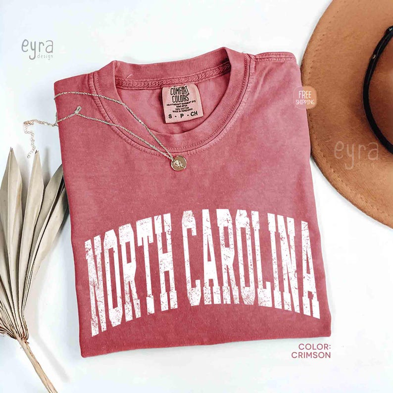 K&ouml;nnte beinhalten: Ein karmesinrotes Kurzarm-T-Shirt mit einem verwaschenen, wei&szlig;en, bogenf&ouml;rmigen Schriftzug, der NORTH CAROLINA lautet. Das Shirt hat einen Rundhalsausschnitt, ein Comfort Colors-Etikett am Innenkragen und eine zarte, goldfarbene Halskette.