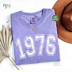 May include: A violet-colored t-shirt with the year "1976" printed in white, distressed lettering. A gold necklace with a pendant rests on the shirt. A brown hat is visible in the background.