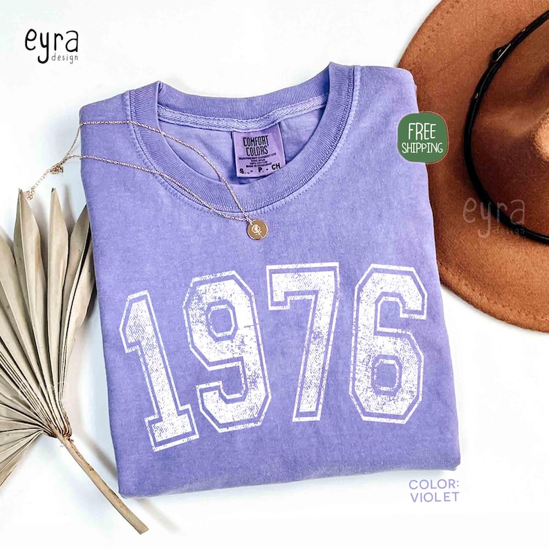 May include: A violet-colored t-shirt with the year "1976" printed in white, distressed lettering. A gold necklace with a pendant rests on the shirt. A brown hat is visible in the background.