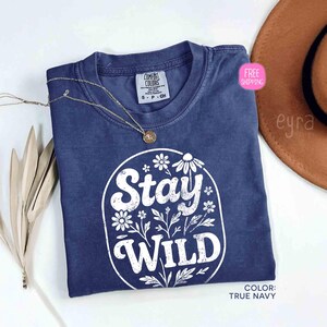 May include: A navy blue t-shirt with the words "Stay Wild" in a white floral design. The shirt has a crew neck and short sleeves. A gold necklace with a pendant is draped across the shirt. The color of the shirt is true navy.