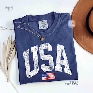 Puede incluir: Una camiseta azul marino con la palabra "USA" en letras blancas grandes y desgastadas. Debajo del texto hay una peque&ntilde;a bandera estadounidense. Un collar dorado descansa sobre la camiseta. El color de la camiseta es "True Navy".