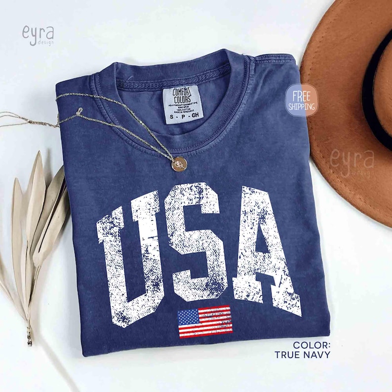 Puede incluir: Una camiseta azul marino con la palabra "USA" en letras blancas grandes y desgastadas. Debajo del texto hay una peque&ntilde;a bandera estadounidense. Un collar dorado descansa sobre la camiseta. El color de la camiseta es "True Navy".