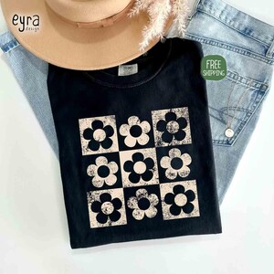 May include: Black t-shirt featuring a retro floral design in a grid pattern. The flowers are in a cream color with black outlines, set against a black background. The shirt is paired with light-wash denim and a tan hat.