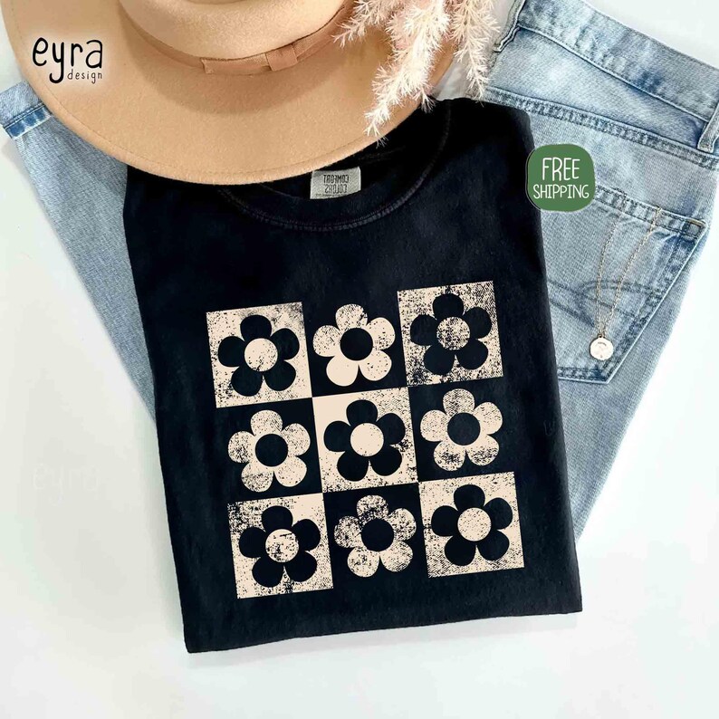 May include: Black t-shirt featuring a retro floral design in a grid pattern. The flowers are in a cream color with black outlines, set against a black background. The shirt is paired with light-wash denim and a tan hat.