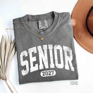 May include: A gray Comfort Colors t-shirt with the word "SENIOR" in large, distressed white letters and the year "2027" below. A delicate silver necklace with a pendant rests on the shirt. The shirt is folded on a white surface.