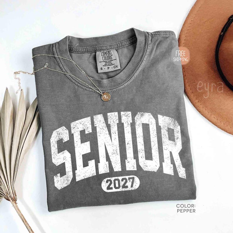 May include: A gray Comfort Colors t-shirt with the word "SENIOR" in large, distressed white letters and the year "2027" below. A delicate silver necklace with a pendant rests on the shirt. The shirt is folded on a white surface.