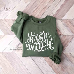 Può includere: Una felpa a maniche lunghe color verde militare con testo stampato in bianco che recita "BASIC WITCH" in un carattere stilizzato, circondato da piccoli elementi grafici tra cui stelle, una luna crescente e due pipistrelli.