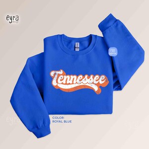 K&ouml;nnte beinhalten: Ein k&ouml;nigsblaues Langarm-Sweatshirt mit Rundhalsausschnitt und dem Wort Tennessee in wei&szlig;er, abgenutzter Schreibschrift mit orangefarbener Umrandung. Das Kleidungsst&uuml;ck hat einen gerippten Kragen, B&uuml;ndchen und Saum sowie ein sichtbares Etikett.