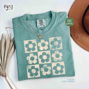 May include: Light green t-shirt with a grid of cream-colored flowers. The shirt has a crew neck and short sleeves. A silver necklace with a pendant rests on the shirt. The tag reads "COMFORT COLORS". The color is "LIGHT GREEN".