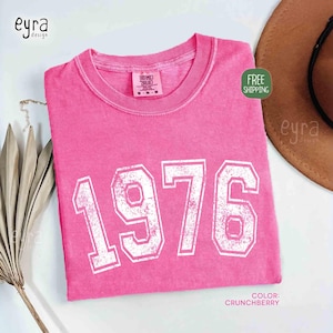 May include: A pink t-shirt with the year "1976" printed in white, distressed block letters. The shirt is a solid color and has a crew neck. The color is described as "Crunchberry".