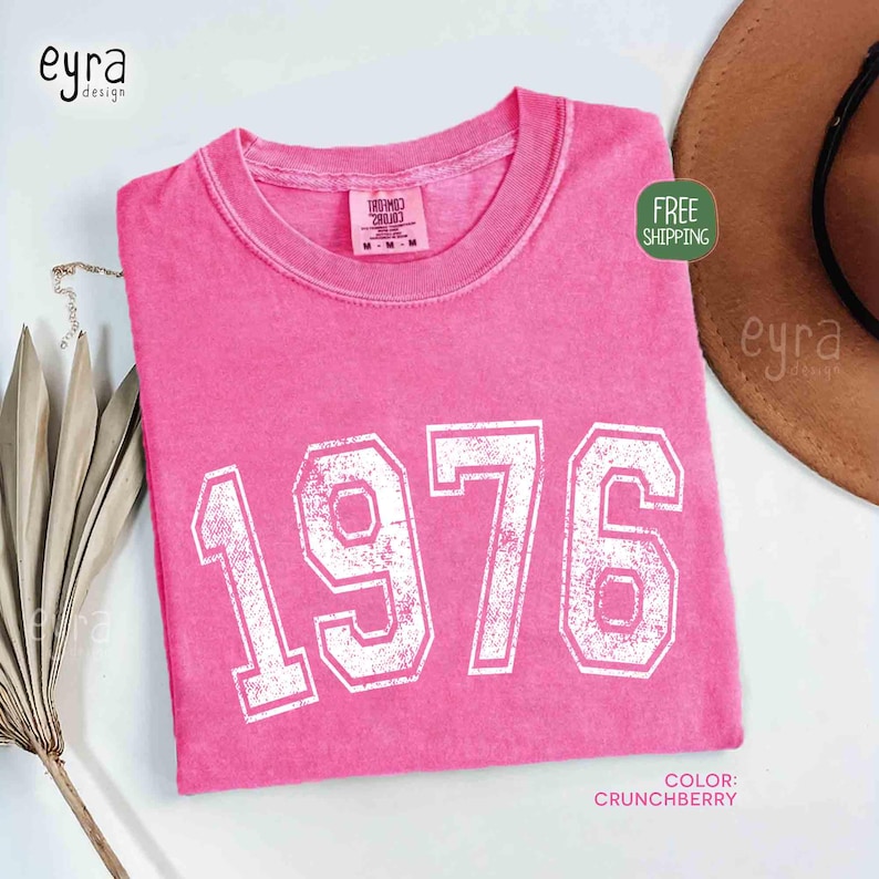 May include: A pink t-shirt with the year "1976" printed in white, distressed block letters. The shirt is a solid color and has a crew neck. The color is described as "Crunchberry".