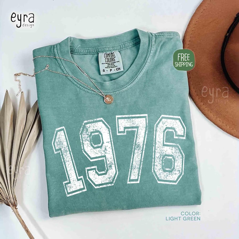 May include: Light green t-shirt with the year "1976" printed in white, featuring a crew neck and a small tag. A gold necklace with a pendant rests on the shirt. The shirt is folded on a white surface.