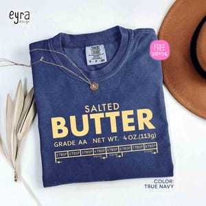 May include: A true navy blue t-shirt with the words "SALTED BUTTER" in large yellow letters. Below the text is "GRADE AA NET WT. 4 OZ. (113g)" and a measuring guide. A gold necklace rests on the shirt.