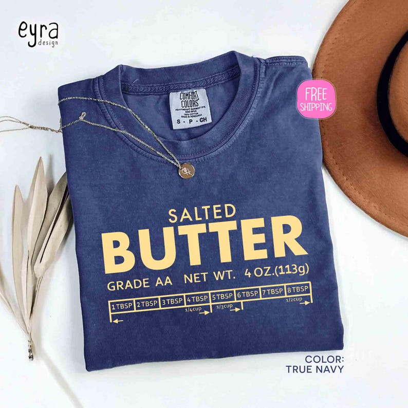 May include: A true navy blue t-shirt with the words "SALTED BUTTER" in large yellow letters. Below the text is "GRADE AA NET WT. 4 OZ. (113g)" and a measuring guide. A gold necklace rests on the shirt.