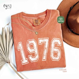 May include: A coral-colored t-shirt with the year "1976" printed in white, vintage-style lettering. A gold necklace and a brown hat are next to the shirt. The shirt has a Comfort Colors tag. The color of the shirt is "Yam".