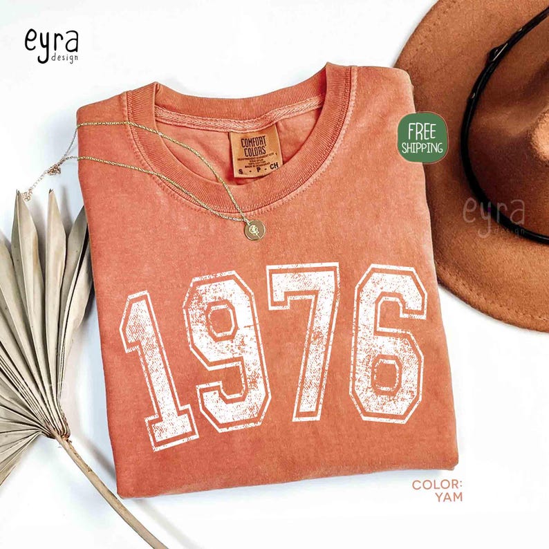 May include: A coral-colored t-shirt with the year "1976" printed in white, vintage-style lettering. A gold necklace and a brown hat are next to the shirt. The shirt has a Comfort Colors tag. The color of the shirt is "Yam".