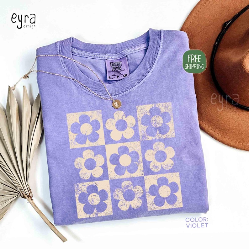 May include: A violet Comfort Colors t-shirt with a grid of beige flowers. A gold necklace with a pendant rests on the shirt. The shirt is folded, with a brown hat in the background. The text "COLOR: VIOLET" is visible.