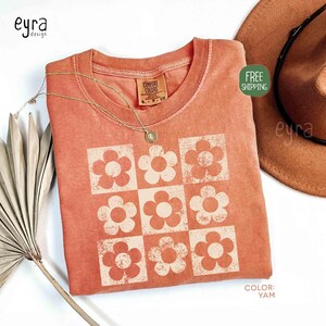 May include: A coral-colored t-shirt with a grid of cream-colored flower designs. The shirt has a crew neck and short sleeves. A gold necklace with a pendant rests on the shirt. A brown hat is visible in the background.