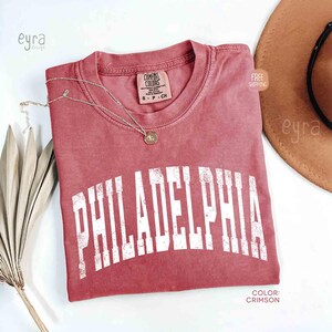 May include: A crimson short-sleeve t-shirt with a distressed white arched graphic that reads PHILADELPHIA across the chest. The shirt features a crew neckline, a Comfort Colors brand label, and a thin gold-toned chain necklace resting on top.