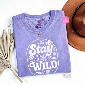 May include: A violet Comfort Colors t-shirt with the words "Stay Wild" in a white floral design. A gold necklace and a brown hat are next to the shirt. The shirt has a crew neck and short sleeves.