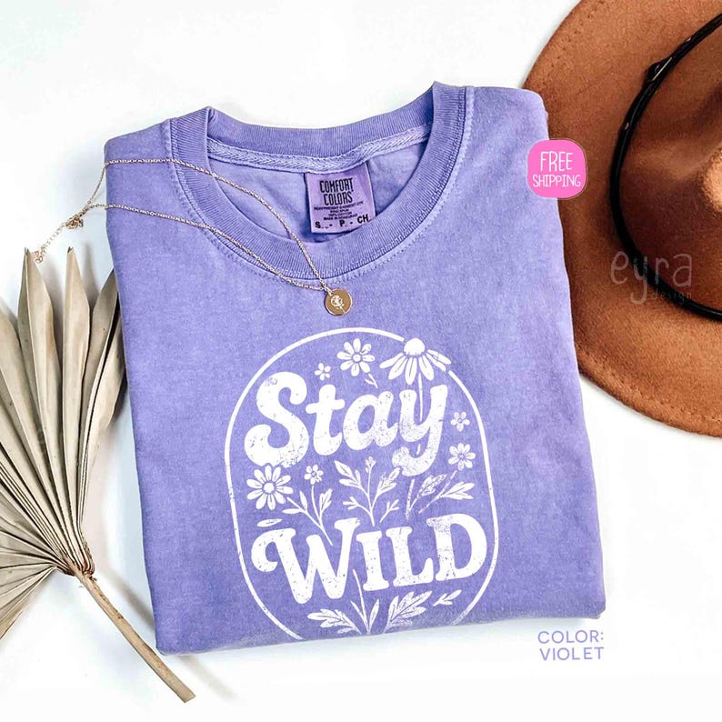 May include: A violet Comfort Colors t-shirt with the words "Stay Wild" in a white floral design. A gold necklace and a brown hat are next to the shirt. The shirt has a crew neck and short sleeves.
