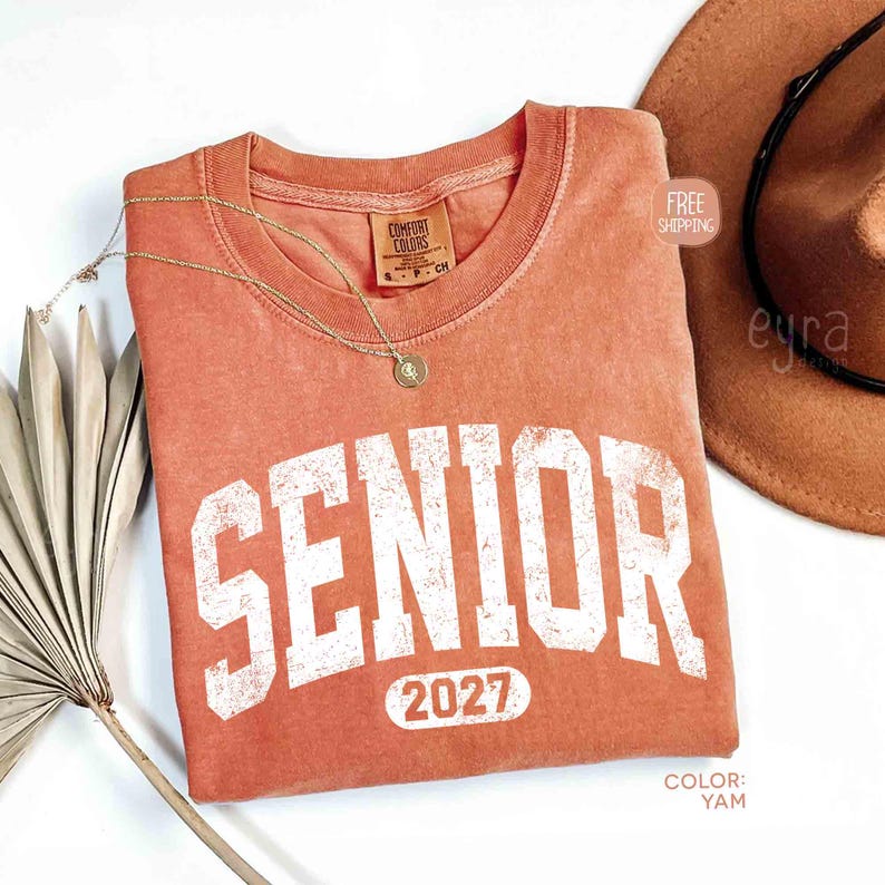 May include: A coral-colored Comfort Colors t-shirt with the word "SENIOR" in large, distressed white letters and the year "2027" below. A gold necklace and a brown hat are also visible.